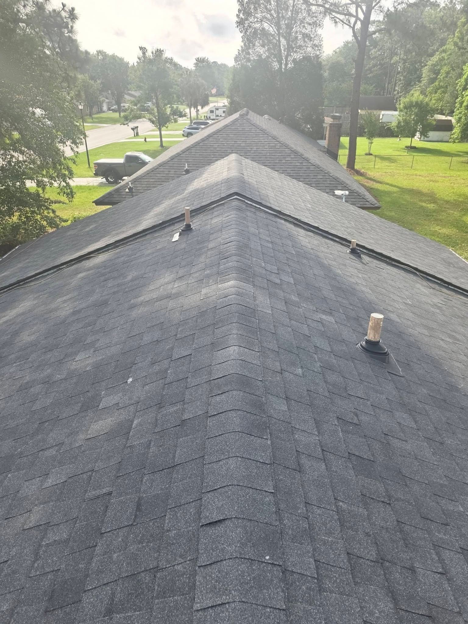 Rooftop with dark shingles viewed along a ridge, bordered by trees and a grassy street below.
