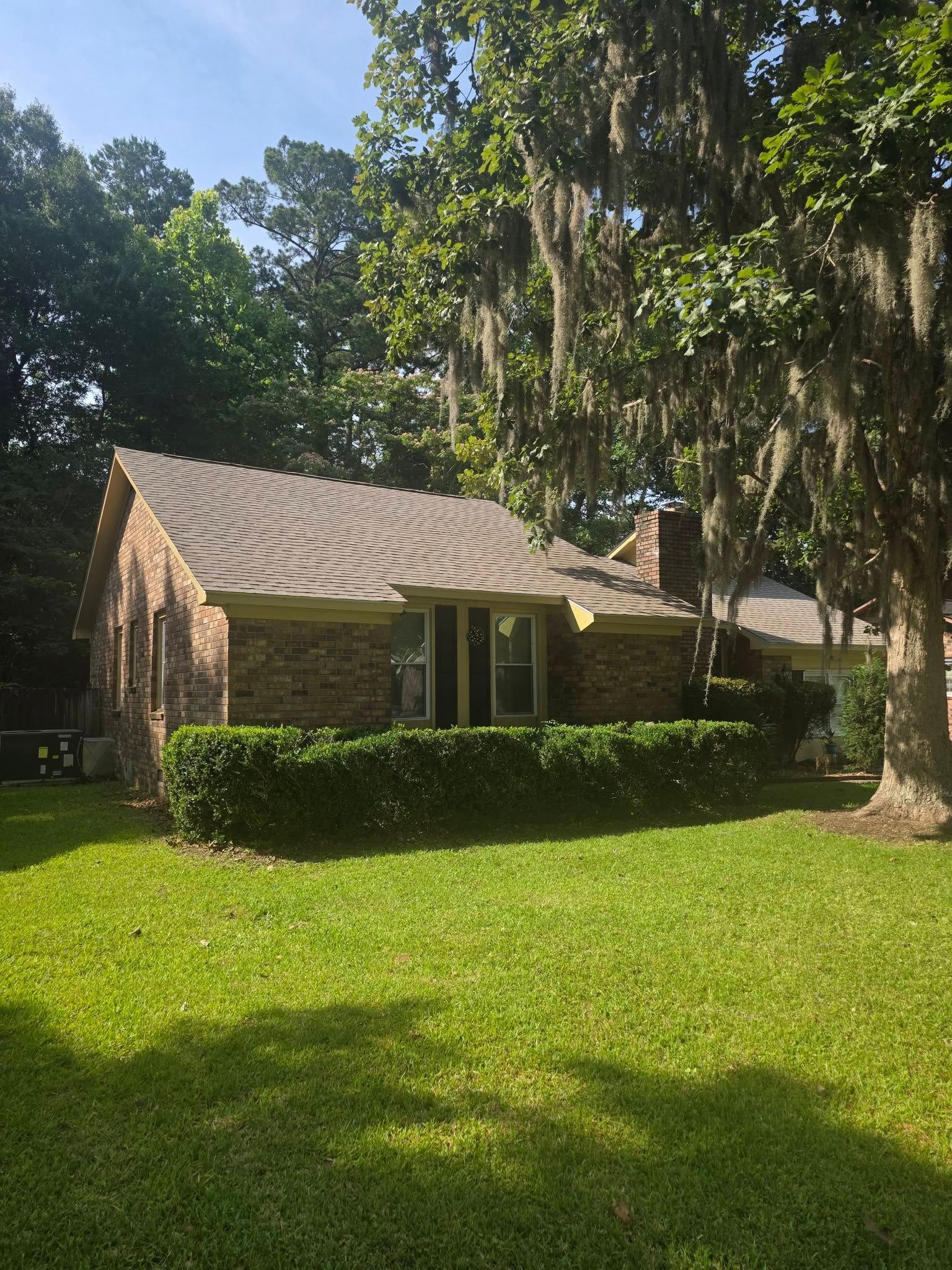 Brick house with a shingled roof and green lawn, surrounded by trees in bright sunlight
