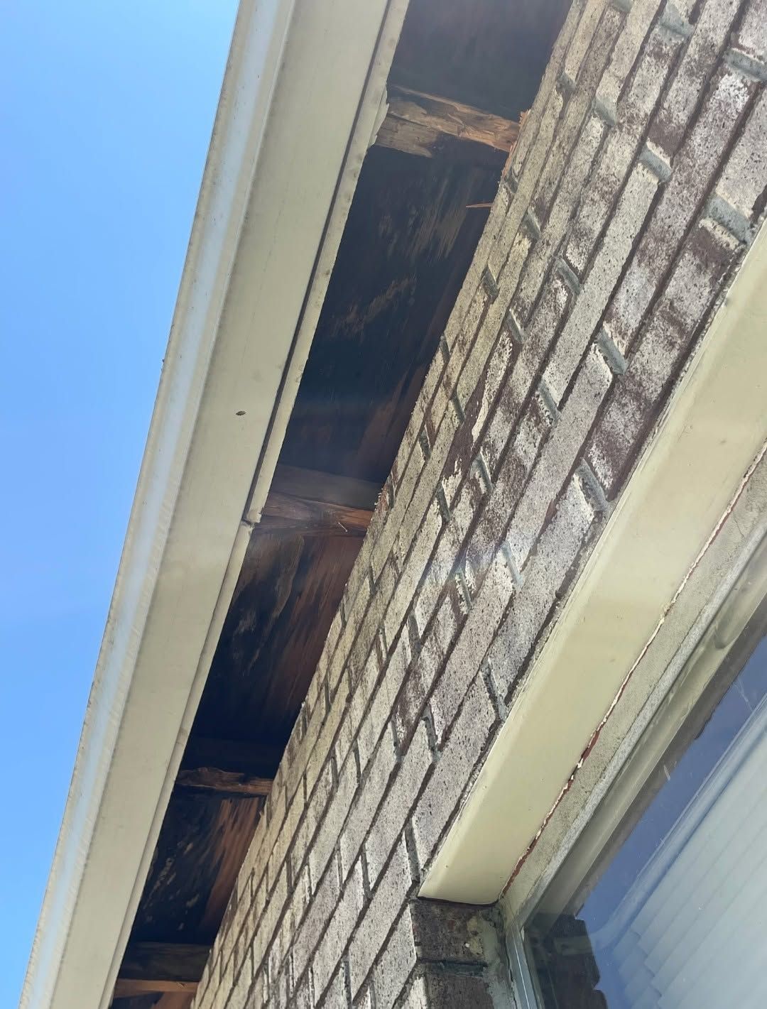 Upward view of damaged roof eaves with exposed wood beside a brick wall and window.