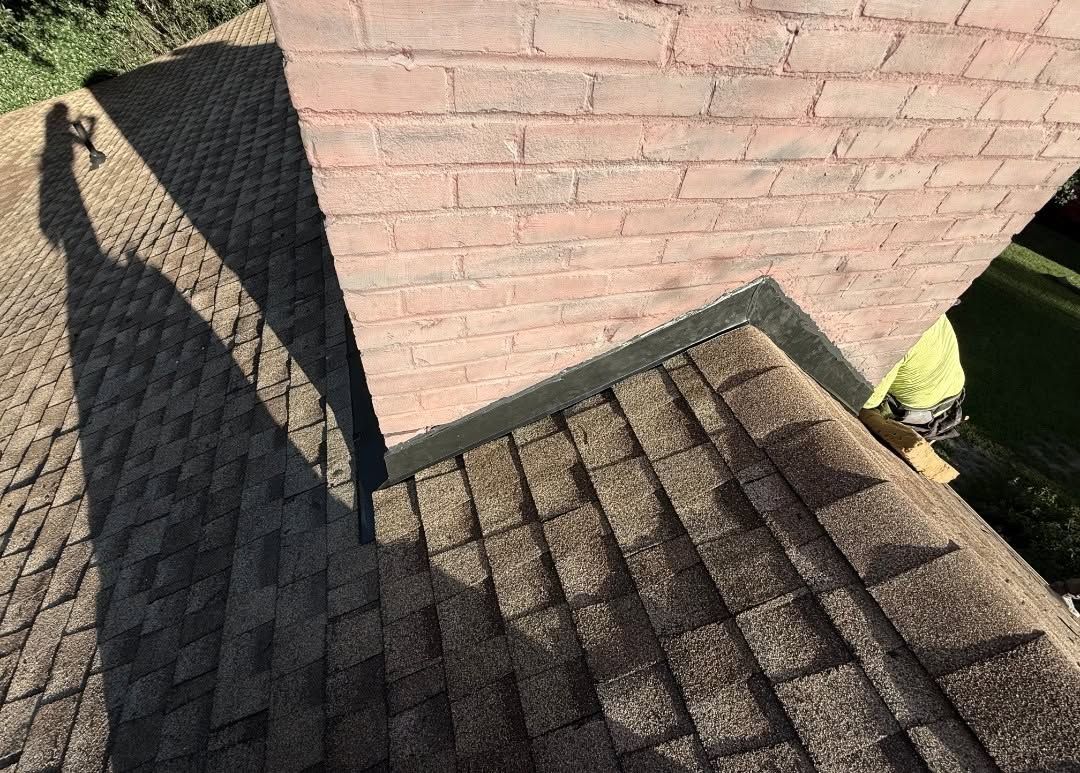 Roof shingles with a large dark shadow cast across them beside a brick wall and gutter.