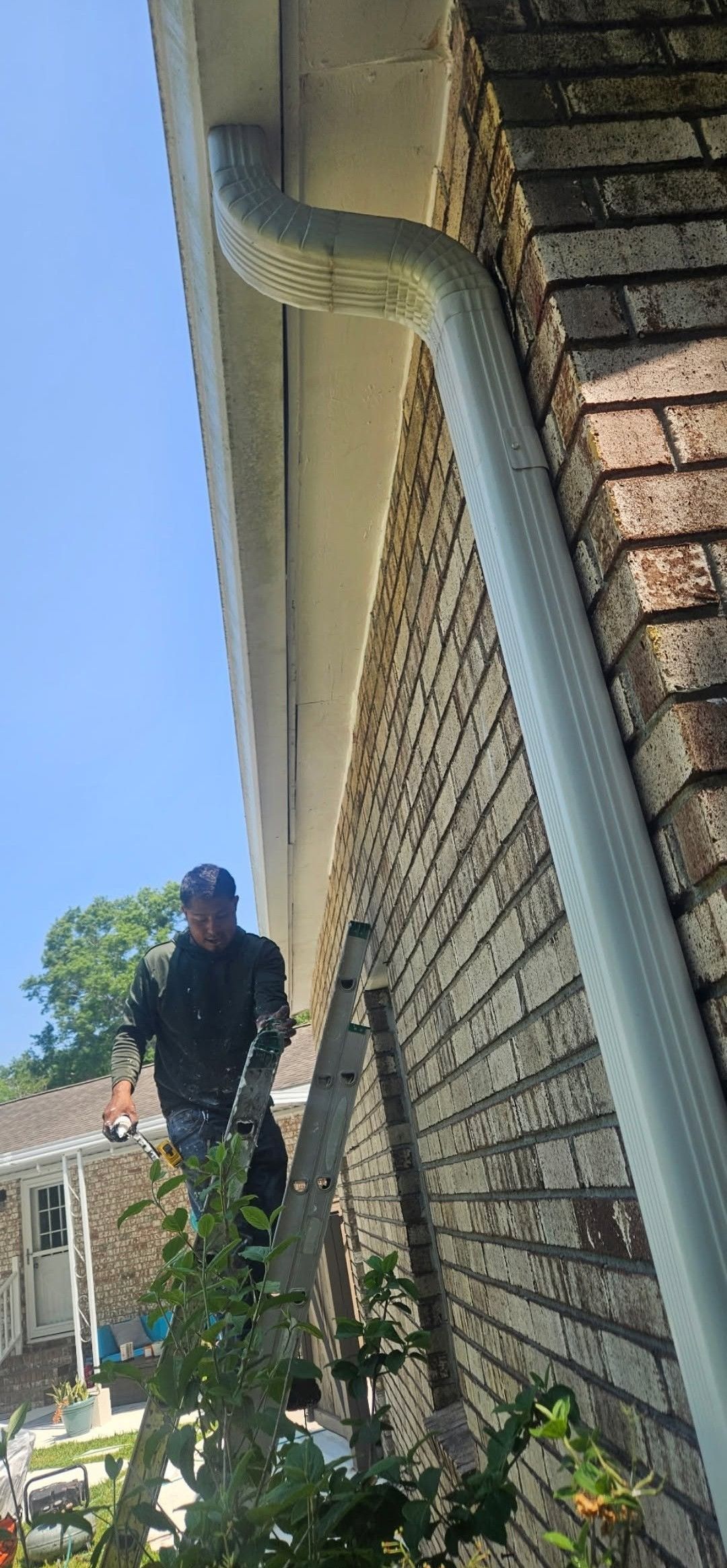 Person standing beside a brick house wall with a white downspout on a sunny day