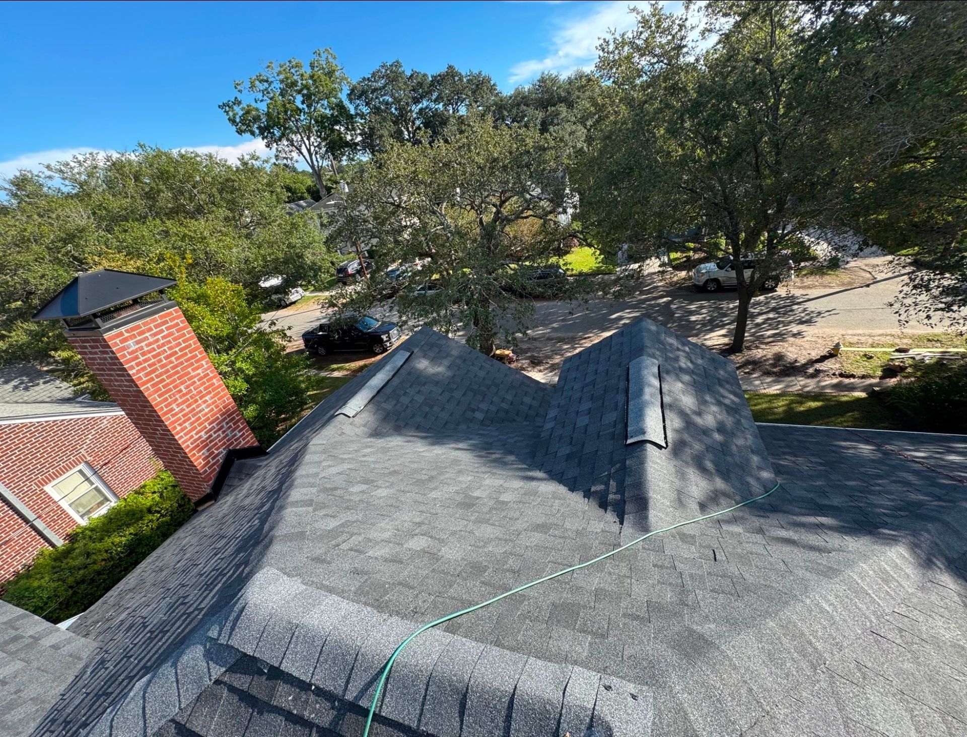 Rooftop with dark shingles beside a red brick chimney, overlooking trees under a blue sky.