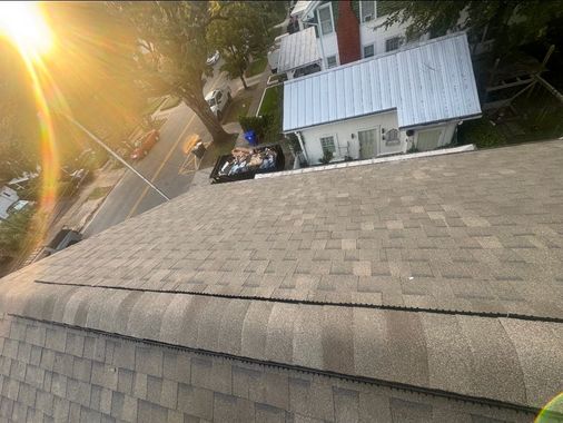 Sunlit rooftop with shingles, overlooking a white house and trees on a steep hillside