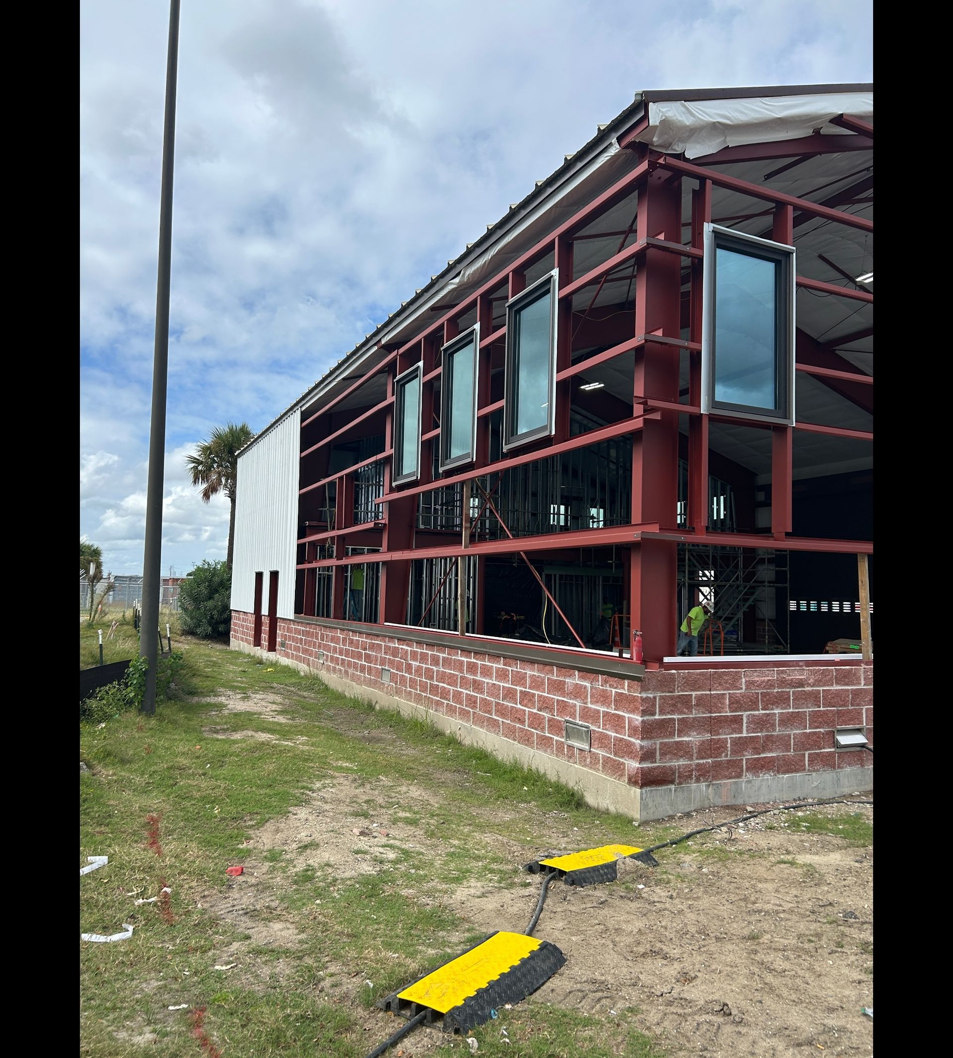 Partially constructed red steel building beside a brick wall and grassy roadside under a cloudy sky