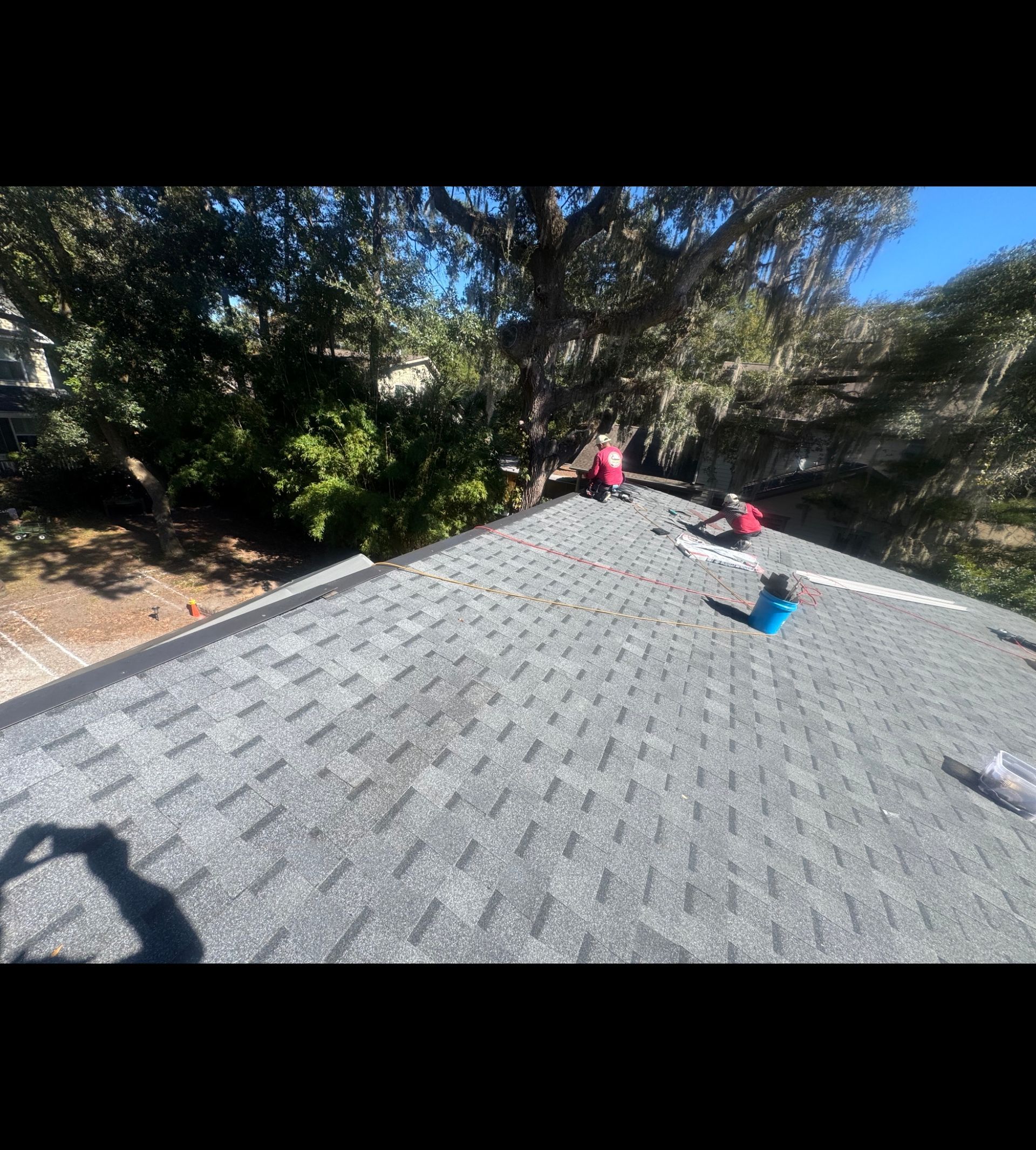 Workers on a gray shingled roof near trees, with one in red kneeling and another in blue nearby.