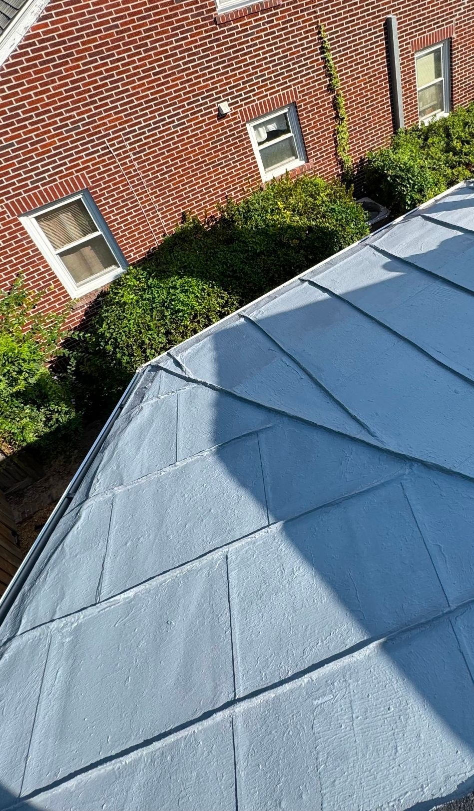 Blue-gray roof in foreground beside a red brick house with small windows and green shrubs below.