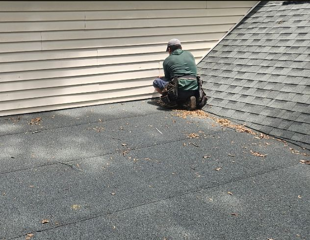 Worker repairing a shingled roof next to beige siding, kneeling beside a wall.