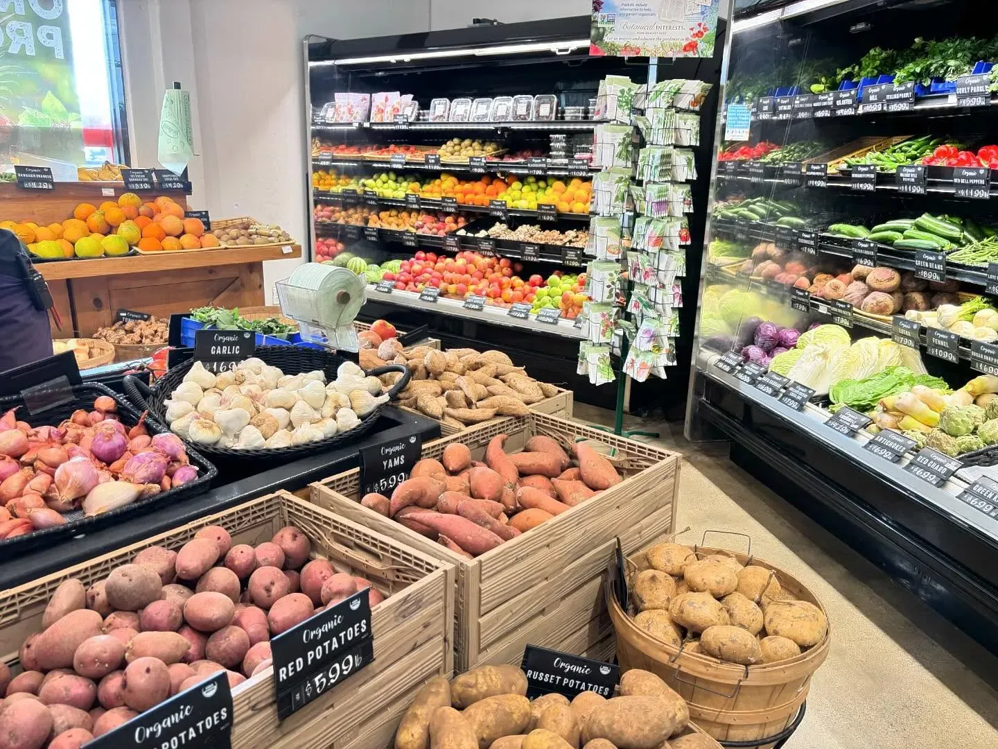 Produce section in a grocery store featuring bins of various potatoes and onions with shelves of fresh produce behind.