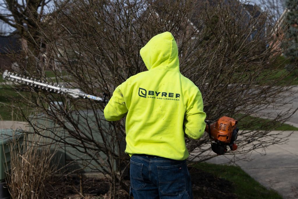 Person in neon yellow hoodie using a gas-powered trimmer near a tree in a yard.