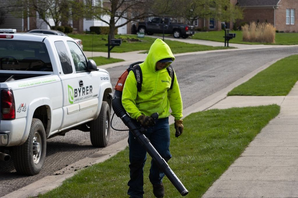 Worker in neon green jacket using a leaf blower on a suburban sidewalk beside a utility truck.