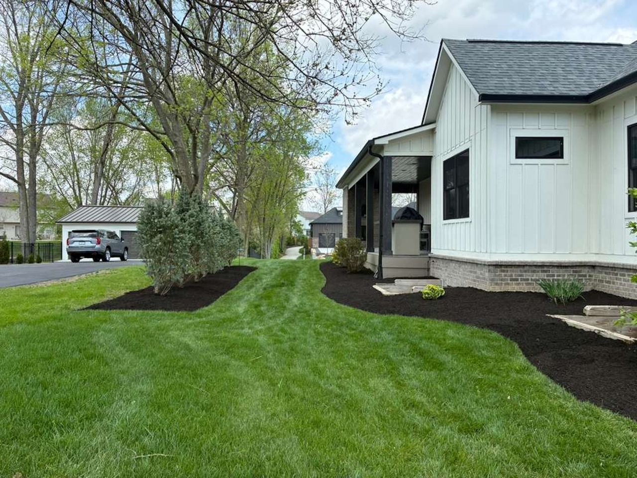 White house with black trim beside a green lawn and landscaped mulch beds under trees.