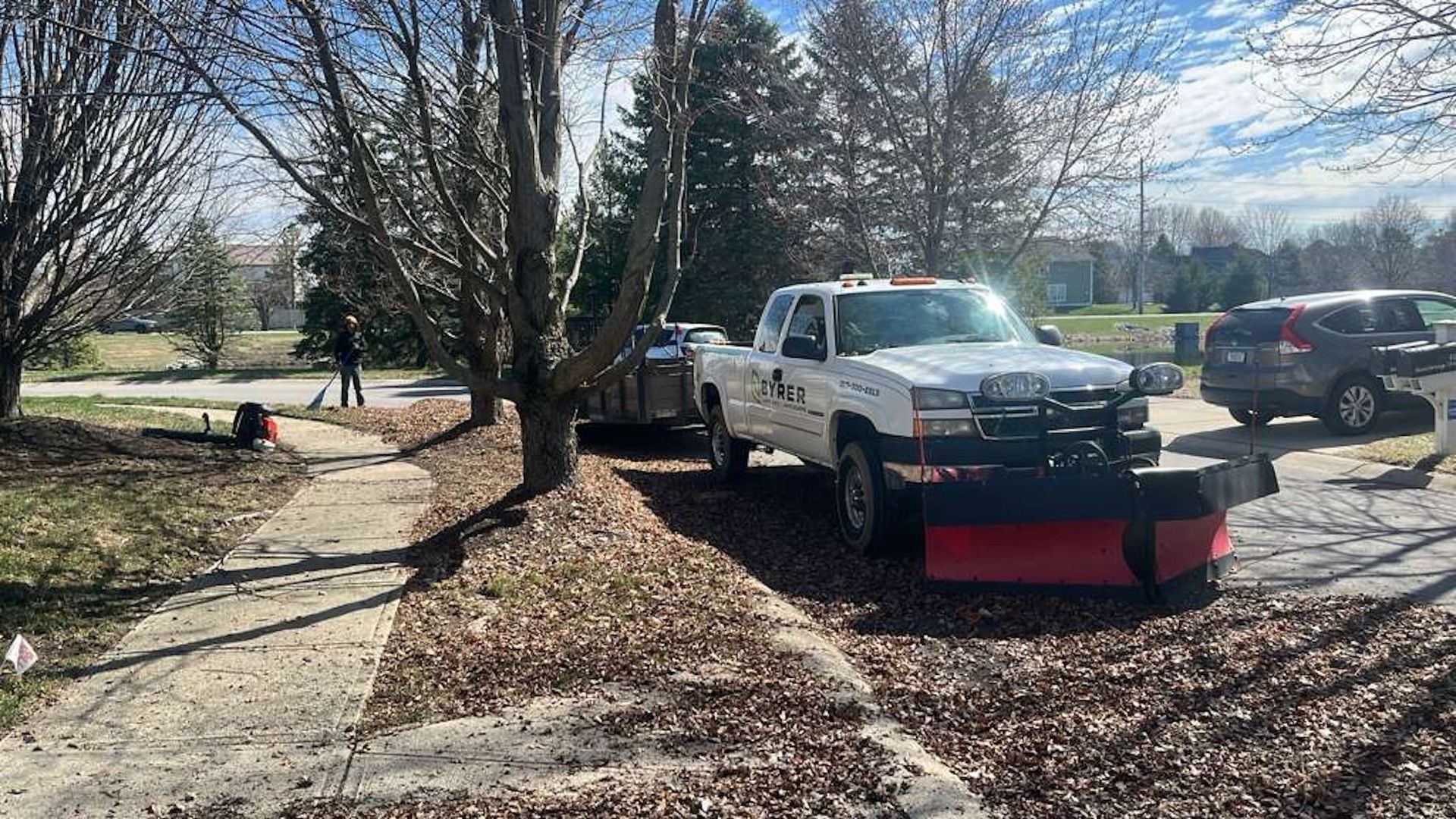 White pickup with red snowplow parked by sidewalk in a leaf-covered yard on a sunny day