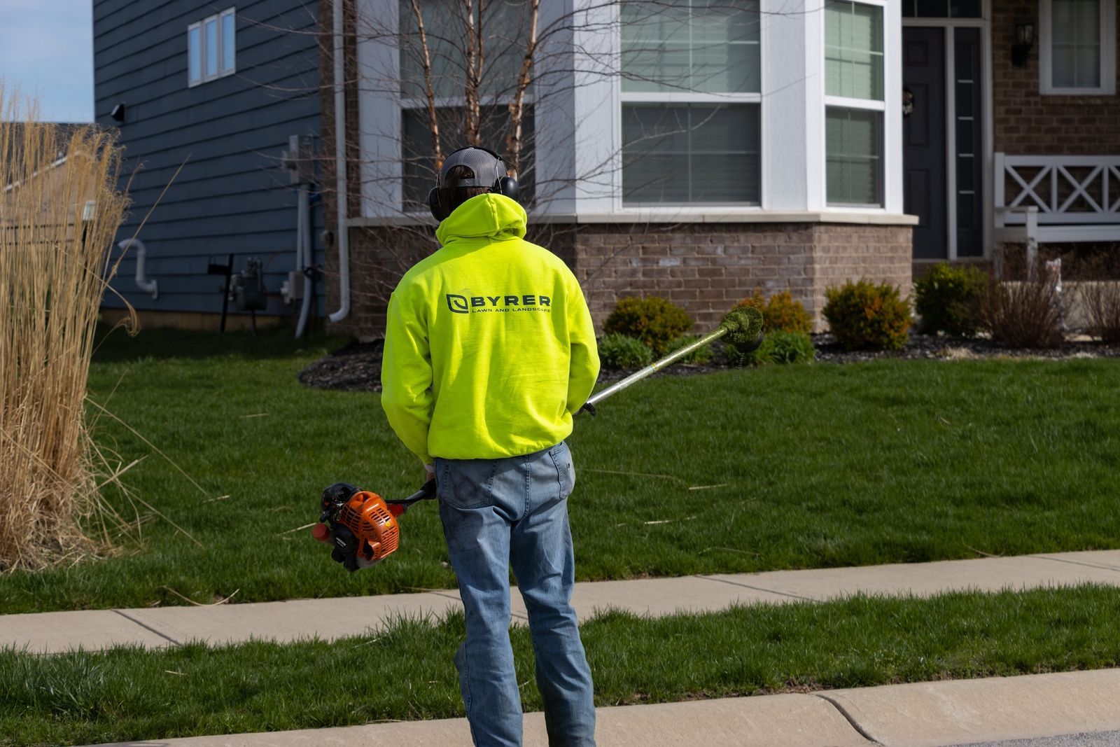 Person in a neon yellow jacket using a leaf blower on a suburban sidewalk.
