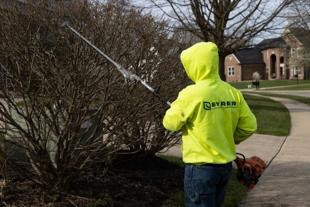 Person in neon yellow jacket using a long pole beside bushes on a sidewalk near a house.