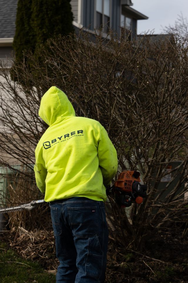Person in a neon yellow hoodie using a hedge trimmer beside a bush in a residential yard