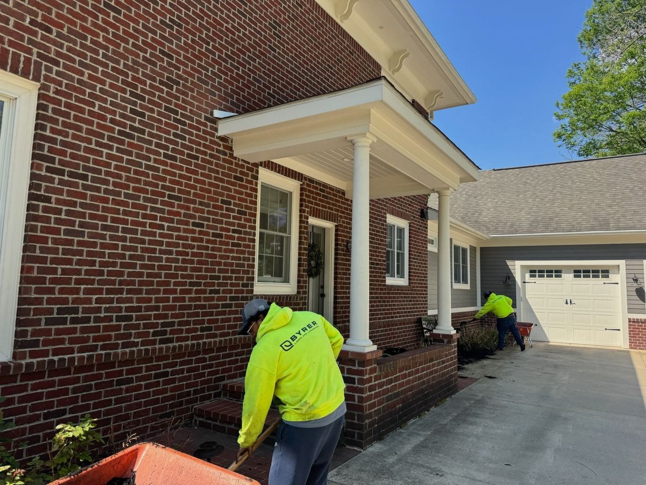 Workers cleaning exterior brick house and driveway, one near the porch and one by the garage.