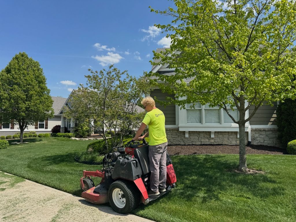 Person mowing a lawn with a riding mower beside a house on a sunny day