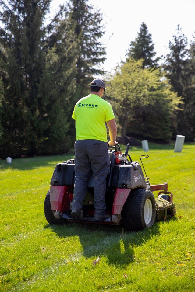 Person mowing a grassy lawn on a riding mower beside trees and shrubs