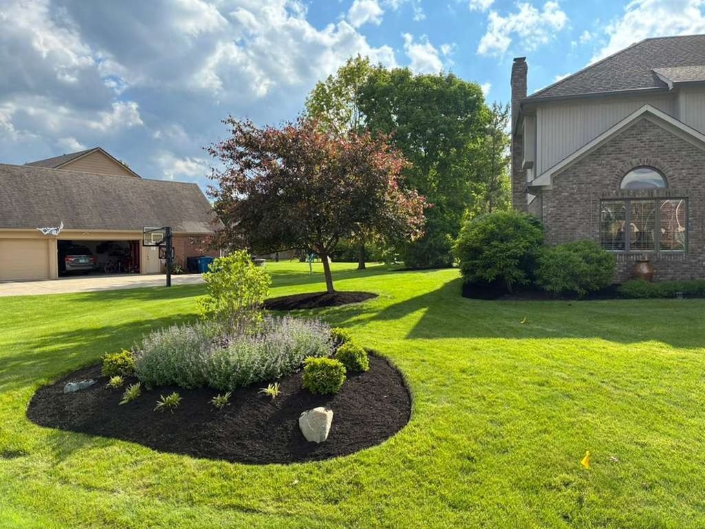 Sunlit suburban front yard with a landscaped flower bed, green lawn, driveway, and brick houses