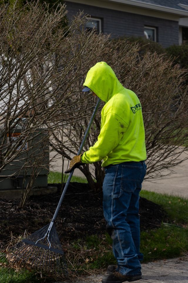 Person in a neon yellow hoodie raking leaves beside a house and bushes