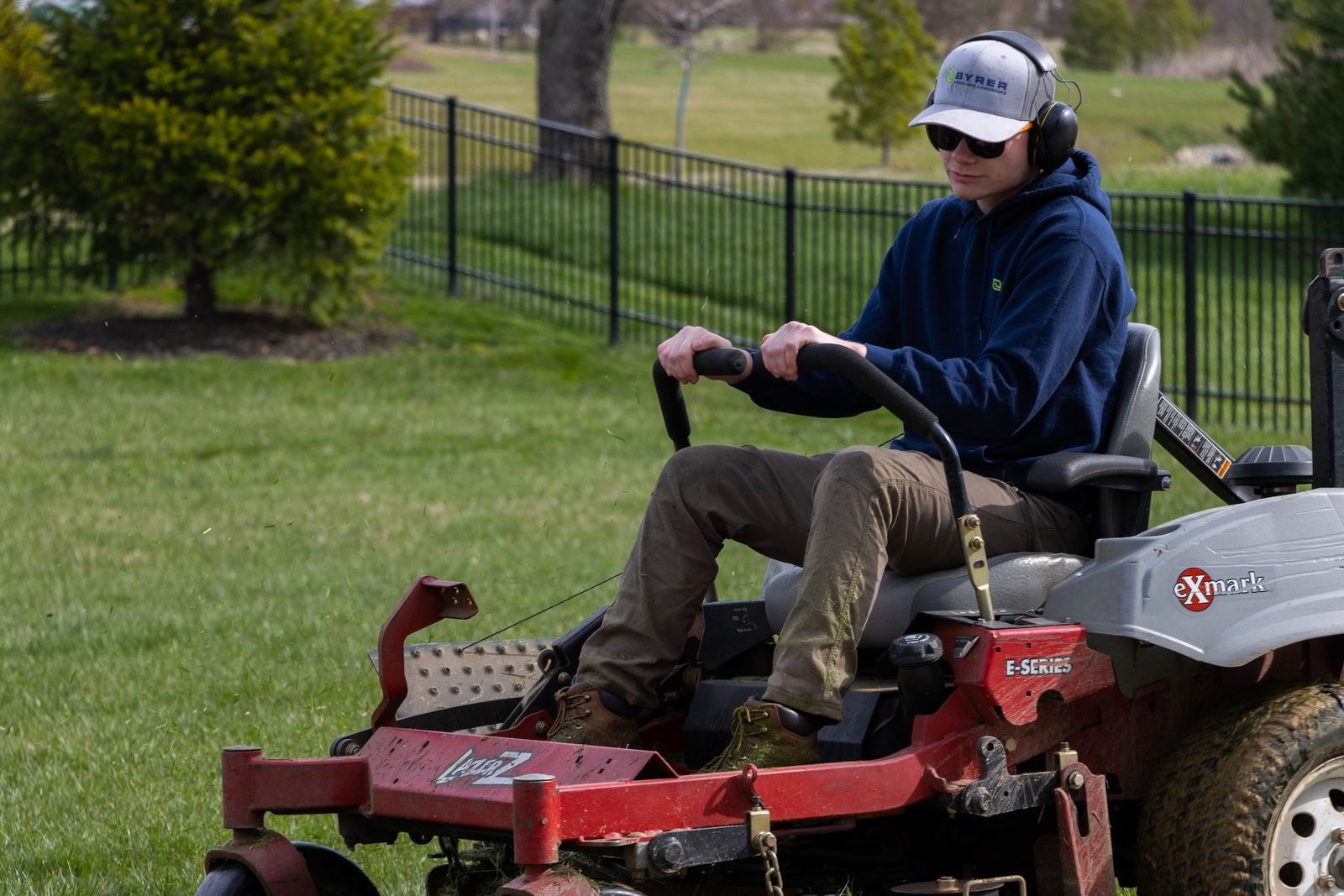 Person riding a red lawn mower on grass, wearing a blue hoodie and cap.