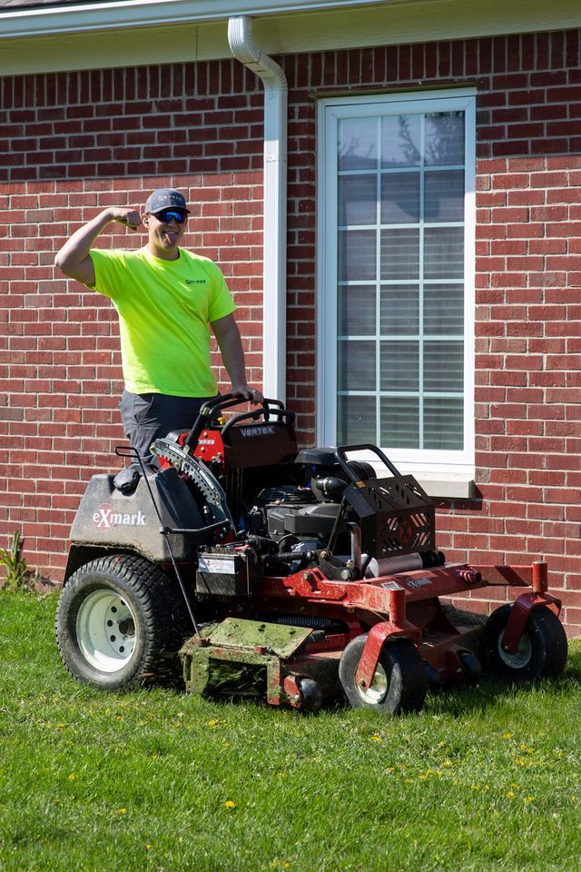 Man in neon shirt saluting on riding mower in front of brick house