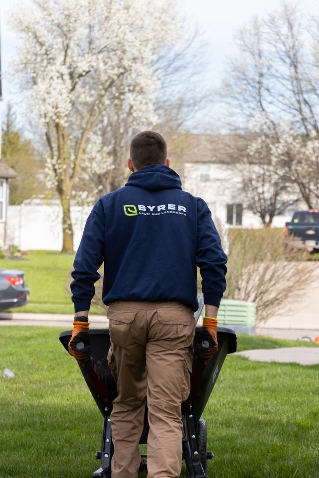 Worker in navy jacket carrying equipment across a grassy yard near houses and flowering trees.