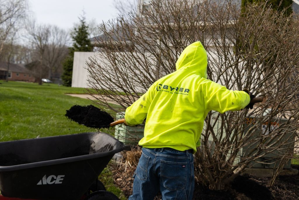 Person in a neon yellow hoodie working in a yard, raking near a wheelbarrow and bushes.