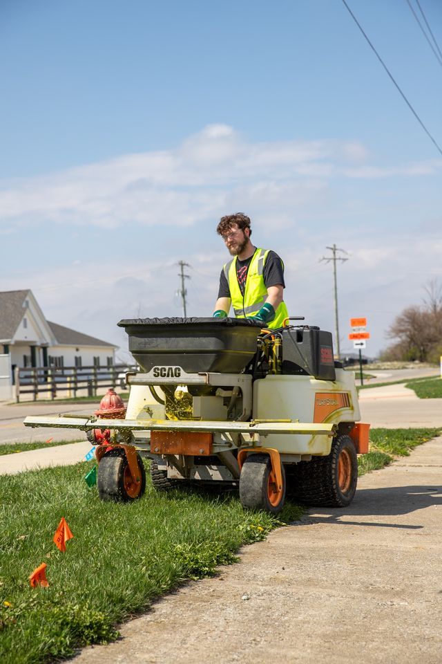 Worker in a neon vest operating a small lawn machine along a sidewalk in a suburban neighborhood