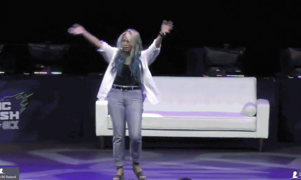 Woman with arms outstretched stands in front of a white couch on a stage.