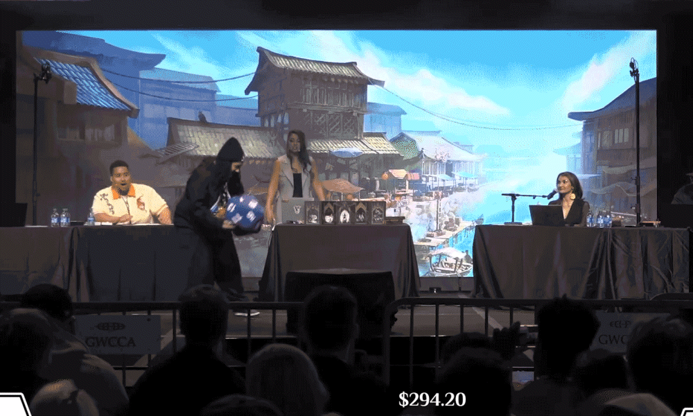 Panel with a presenter holding an object on stage in front of a backdrop. Two people sit on either side.