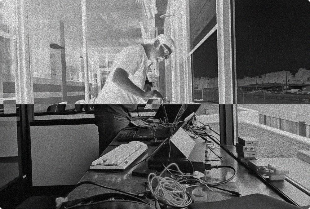 A person works at a desk with electronics, illuminated by a window. Black and white photograph.