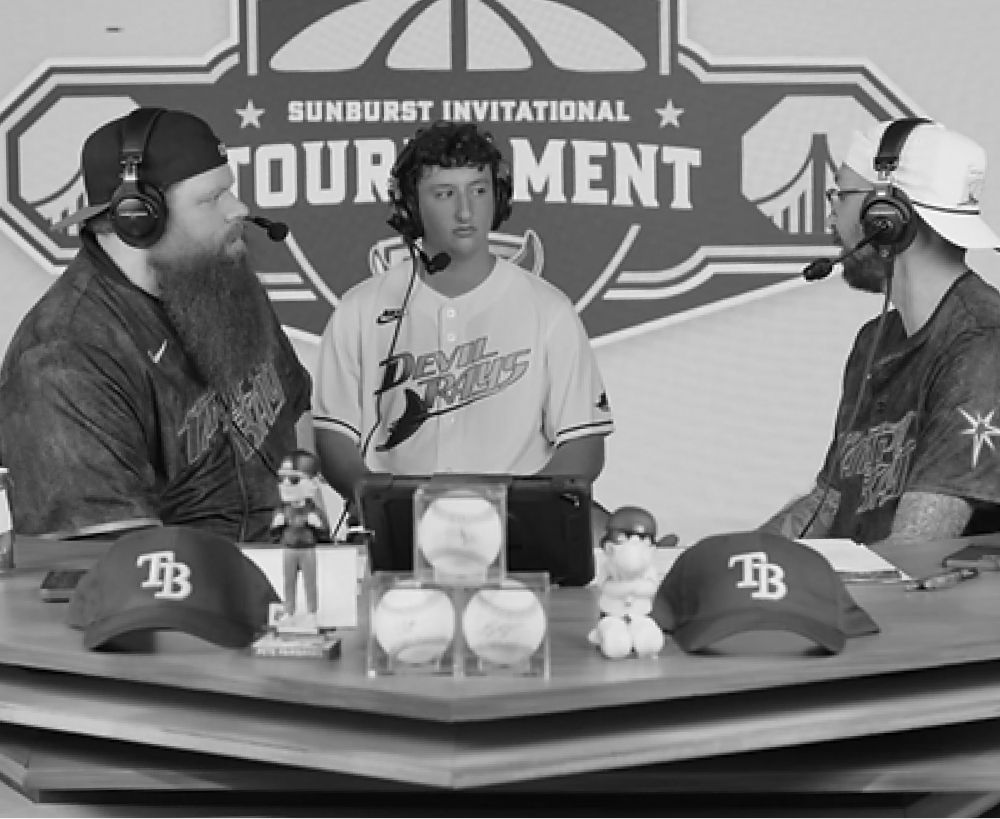 Three people at a baseball tournament table. Two wearing headsets interview a player wearing a baseball jersey.