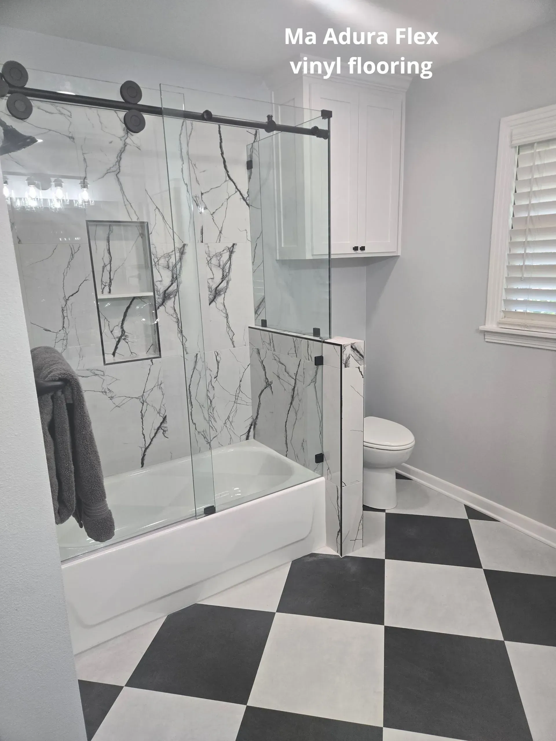 Small bathroom with marble shower, white vanity, and black-and-white checkered vinyl flooring.