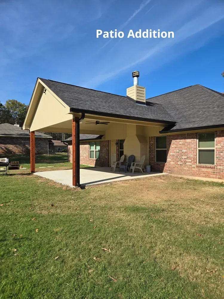 Covered patio addition with brick house, dark roof, and patio chairs in a grassy backyard