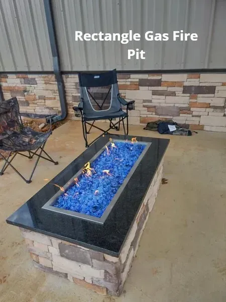 Outdoor rectangular gas fire pit with blue flames beside patio chairs and stone wall.