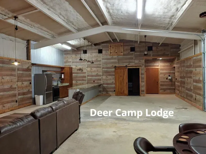 Rustic lodge interior with leather sofa, wood-paneled walls, and doorways; “Deer Camp Lodge” sign on the floor