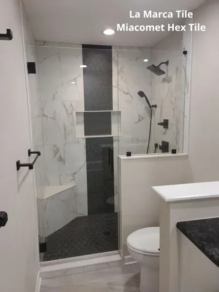 Small bathroom with glass shower, white marble-look tile, black fixtures, and toilet beside a vanity.