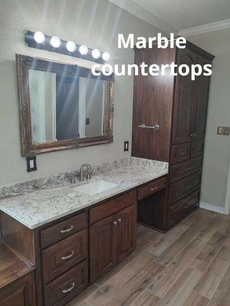 Bathroom vanity with white marble countertop, wood cabinets, mirror, lights, and wood-look floor.