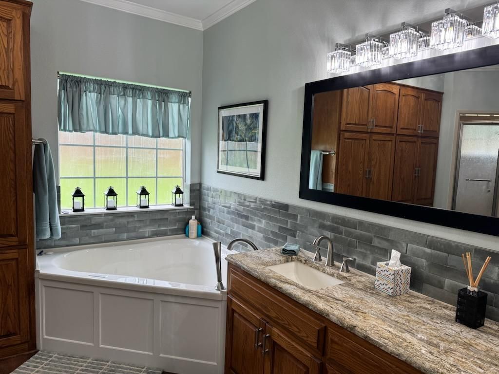 Bright bathroom with gray walls, corner tub, wooden vanity, large mirror, and window over the sink.