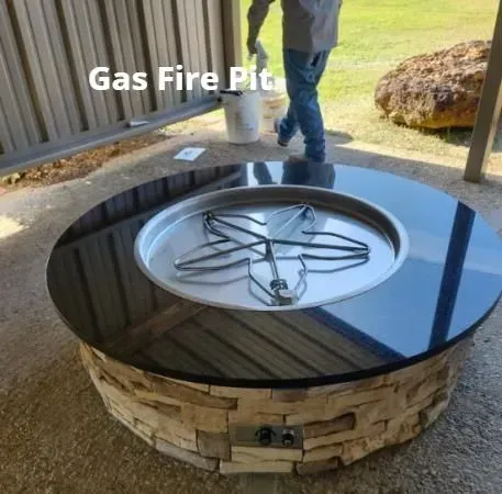 Outdoor gas fire pit with black glass top and stone base on a patio; a person stands nearby.