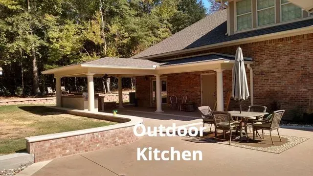 Patio outdoor kitchen with covered seating, brick house backdrop, and grill area in a sunny backyard