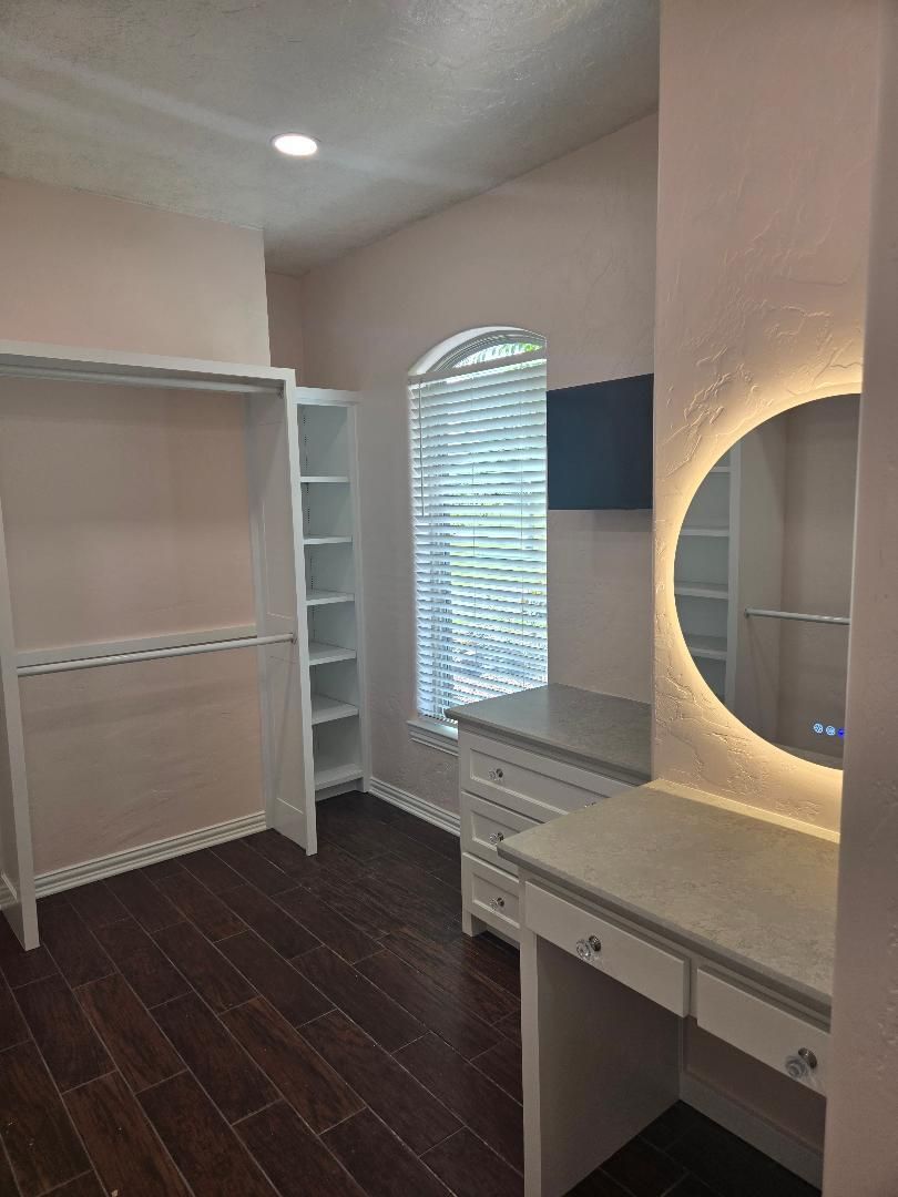 Empty walk-in closet with white shelves, dark wood floor, arched window, and illuminated vanity mirror.