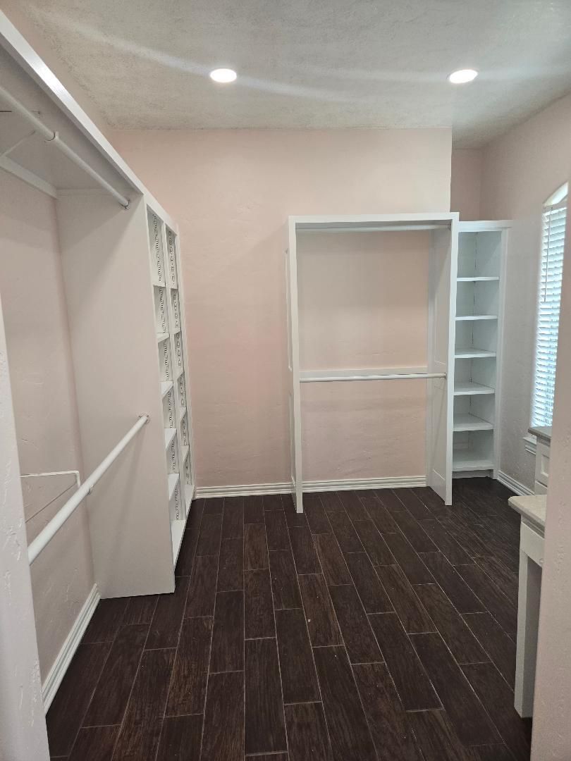 Empty walk-in closet with white shelving and dark wood flooring, viewed toward a windowed wall.