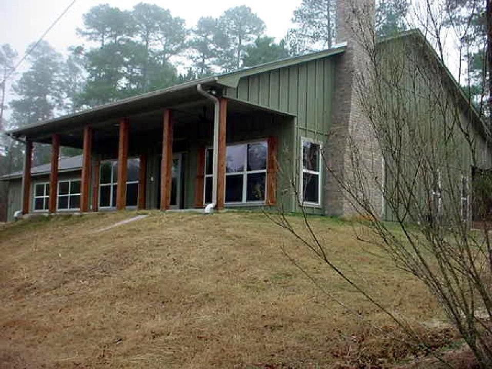 Wooden house on a grassy hill with a covered porch and tall trees behind it