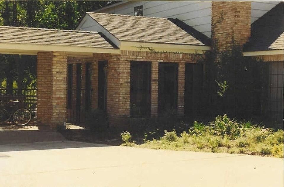Brick house entrance with a covered porch, dark windows, and a sunlit driveway in front.