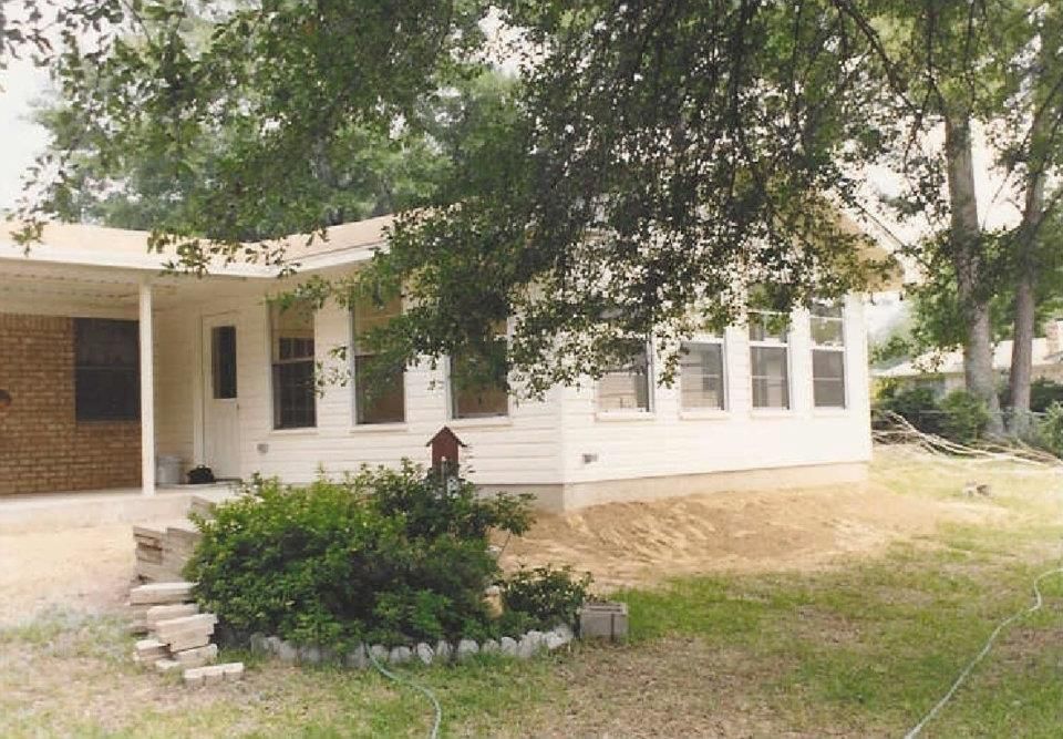 Single-story white house with brick porch, large trees, and a landscaped front yard