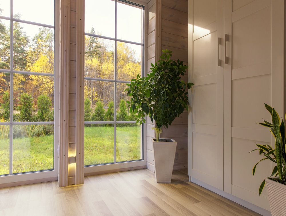 Bright modern room with large windows, light wood floors, and potted plants overlooking a green yard