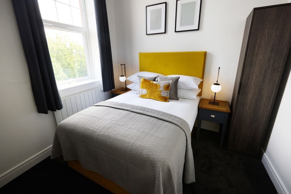 Small bedroom with a bed, yellow headboard, bedside lamps, and a window with dark curtains.