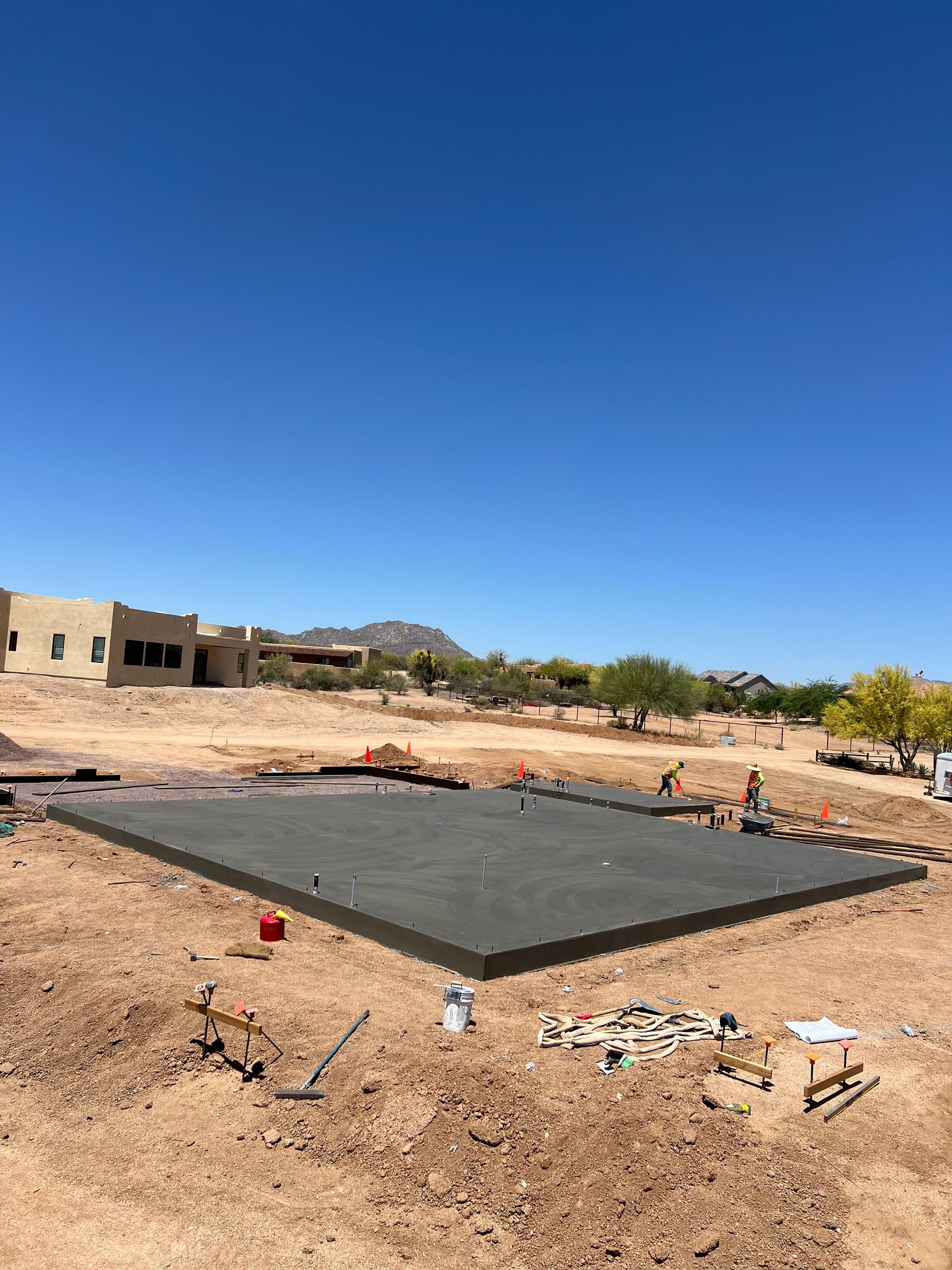 Desert construction site with a large black foundation slab, scattered debris, and a clear blue sky.